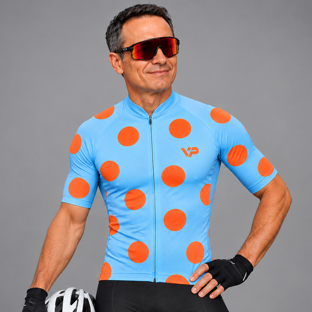 A man modeling a light blue short-sleeve cycling jersey with large orange polka dots and a small orange VP logo, paired with black cycling shorts, black fingerless gloves, and sunglasses, holding a white and black cycling helmet against a solid gray studio background.