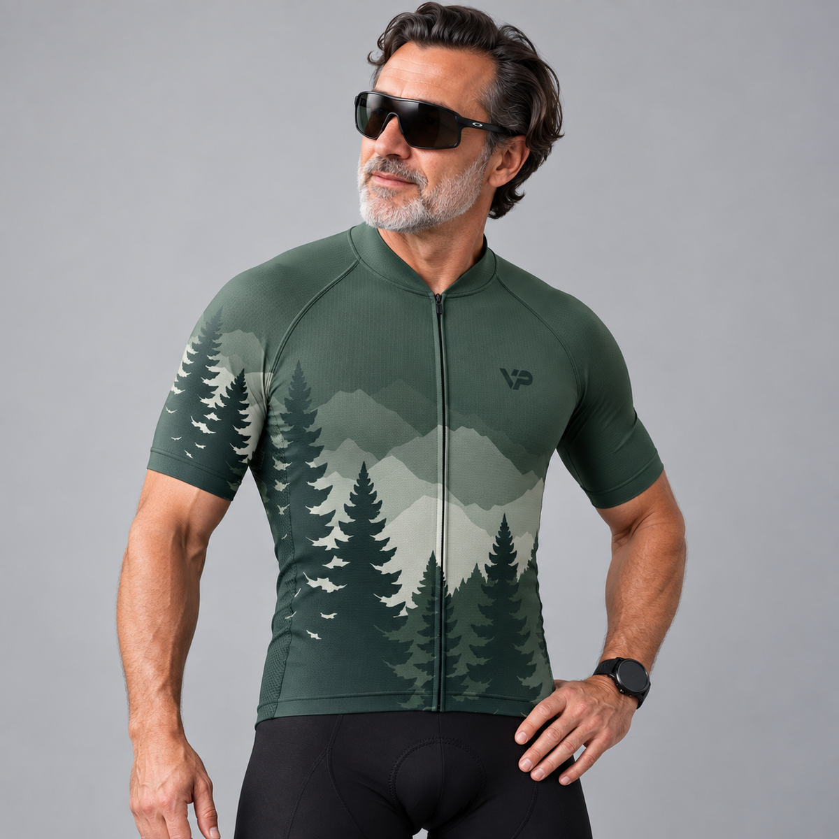 A man wearing a dark green short-sleeve cycling jersey with a pine tree and mountain landscape print, black cycling shorts, and black sunglasses, posed against a solid gray studio background.