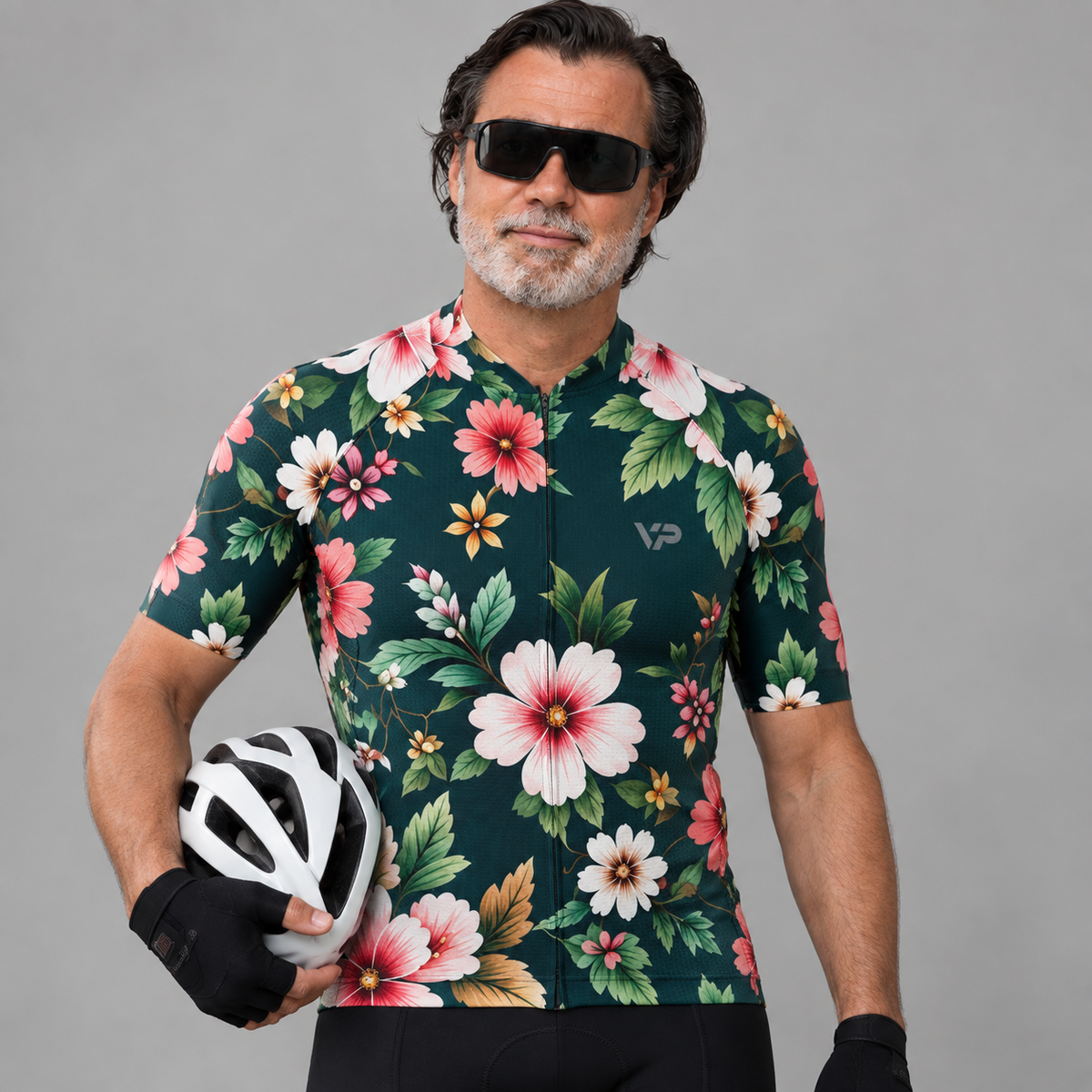 A man wearing a dark green short-sleeve cycling jersey with a vibrant pink, red, and white tropical floral print, holding a white helmet. He is wearing black sunglasses and black cycling shorts against a solid gray studio background.